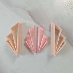 Könnte beinhalten: Art Deco-Stil Keksausstecher und Fondantdekorationen. Die Ausstecher sind hellpfirsichfarben, während die Fondantstücke schimmernd rosa sind. Das Design zeigt eine fächerartige Form, ideal zum Backen und Basteln.