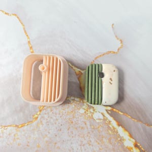 Peut inclure: Un emporte-pièce en plastique pêche avec un cadre rectangulaire et des lattes verticales, placé à côté d'un pendentif rectangulaire présentant une texture côtelée verte d'un côté et une finition marbrée crème lisse de l'autre côté.