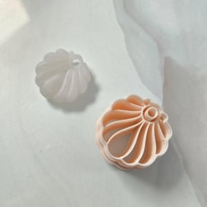 Peut inclure: Un objet blanc, semi-translucide, arrondi et cannelé avec une indentation circulaire centrale, placé à côté d'un emporte-pièce circulaire creux, cannelé et de couleur pêche clair avec un anneau central, présentant un motif radial en coquille.