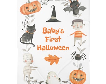 Puede incluir: Una manta de bebé blanca con temática de Halloween. Presenta un gato negro, un sombrero de bruja, una calabaza, un fantasma, una momia, una araña, un Frankenstein y un murciélago. El texto "Baby's First Halloween" está en el centro de la manta.