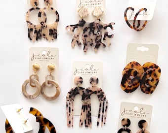 May include: A selection of earrings in neutral colours, featuring tortoiseshell patterns and abstract designs. The earrings are presented on white cards with the text "j.cole CLAY JEWELRY Made in Texas".