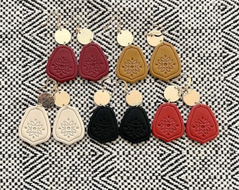 May include: Five pairs of dangle earrings featuring gold-tone circular studs attached to shield-shaped pendants. The pendants come in red, mustard yellow, cream, and black, each embossed with a central floral pattern and metal hook wires.