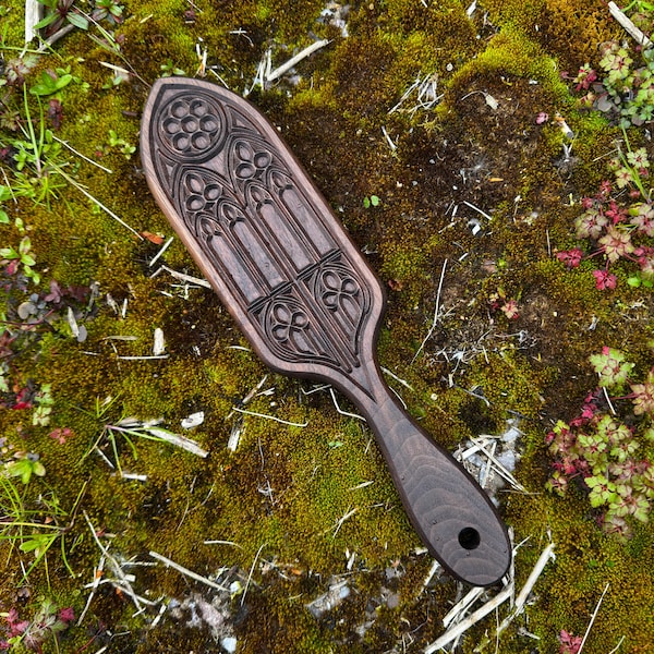 May include: A dark brown wooden paddle with an ornate gothic window design carved into the surface. The paddle has a handle with a hole at the end. The paddle is approximately 12 inches long.