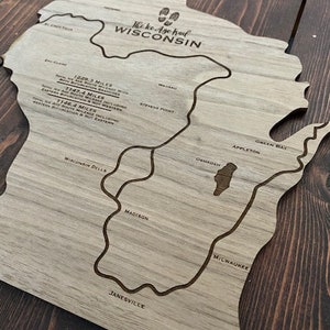 May include: Wooden map of Wisconsin with engraved lines and text. The text includes "The Ice Age Trail WISCONSIN" and city names like Madison and Milwaukee. The map is on a dark wood surface.