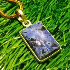 May include: A rectangular pendant with a blue and black stone set in a gold-colored metal frame. The pendant hangs from a gold-colored chain, resting on a bed of green grass. The stone has a marbled appearance.