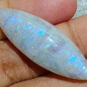 May include: A polished, elongated gemstone with a rounded tip. The stone has a pearly white base color with iridescent blue and purple flashes. The gemstone is smooth and appears to be held in a hand.