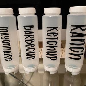 May include: Four clear plastic squeeze bottles with white lids, labeled in black with the words "Mayonnaise", "Barbecue", "Ketchup", and "Ranch". The bottles are designed for dispensing condiments.