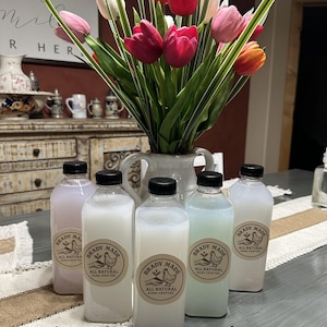 Peut inclure: Six bouteilles transparentes avec des bouchons noirs, étiquetées "Brady Made All Natural Hand Crafted", contenant des liquides aux couleurs pastel. Un vase de tulipes colorées est derrière les bouteilles, sur une table.