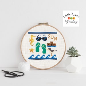 Op de afbeelding: Een witte kruissteek borduurring met een strandthema. Het ontwerp bevat een zeepaardje, een zonnebril, slippers, een ster, een zandkasteel, een meeuw en golven. De tekst "SUN" is geborduurd in gele draad.