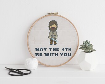 Può includere: Un cerchio da ricamo beige con un disegno a punto croce di un personaggio dei cartoni animati che tiene una spada laser. Il testo "MAY THE 4TH BE WITH YOU" è ricamato sotto il personaggio.