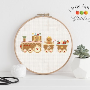 Puede incluir: Un patrón de punto de cruz de un tren de jengibre con dos vagones. El tren está decorado con bastones de caramelo, corazones y un copo de nieve. El patrón está en un aro de bordado blanco.