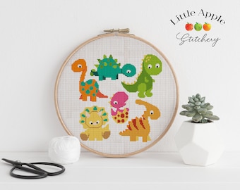 Op de afbeelding: Een kruissteekpatroon met zes kleurrijke dinosaurussen op een witte achtergrond. Het patroon is ingelijst in een houten hoepel en de tekst "Little Apple Stitchery" is zichtbaar in de rechterbovenhoek.