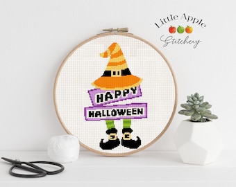 May include: Cross stitch pattern of a witch with a purple banner that says "Happy Halloween". The witch is wearing a black hat with an orange brim and green legs. The pattern is in a wooden embroidery hoop.