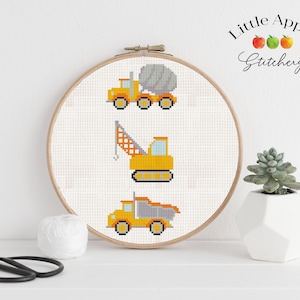 May include: A white embroidery hoop with three cross-stitch designs of construction vehicles. The vehicles are a cement mixer, a crane, and a dump truck. The hoop is on a white surface with a ball of white yarn and a pair of black scissors. The text "Little Apple Stitchery" is in the upper right corner of the image.