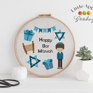 May include: A cross-stitch pattern featuring a boy in a blue shirt and kippah, a Star of David, a Torah scroll, and gifts. The text "Happy Bar Mitzvah" is included in the design. The pattern is displayed in a wooden embroidery hoop.