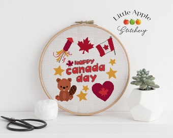 May include: A white embroidery hoop with a cross-stitch design featuring a beaver, a Canadian flag, fireworks, maple leaves, and the text "Happy Canada Day".