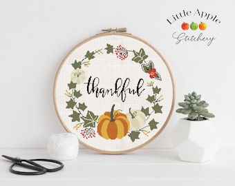 May include: A white embroidery hoop with a cross-stitch design of a wreath with pumpkins, leaves, and berries. The wreath surrounds the word "thankful" in black script. The hoop is on a white surface with a ball of white yarn and black scissors.