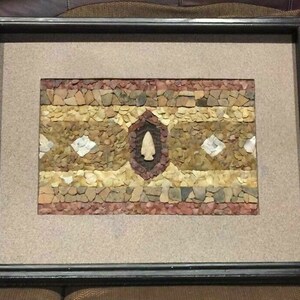 May include: A framed artwork featuring a mosaic design made of small stones and a central arrowhead. The mosaic is in shades of brown, tan, and red, with a diamond pattern.
