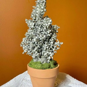 May include: A silver metal tree sculpture in a terracotta pot with green burlap around the base. The tree is made of many small, interconnected pieces that resemble a honeycomb.