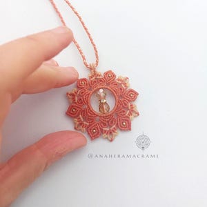 Puede incluir: Un collar de macramé hecho a mano con un colgante en forma de flor de color coral y una cuenta de cristal central. El colgante está adornado con pequeñas cuentas doradas. El collar se muestra sobre una superficie blanca.