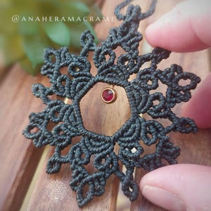 Peut inclure: Un collier en macramé noir fait à la main avec un motif ajouré circulaire. Le collier comporte un petit pendentif rond en pierre rouge. Le texte "@anaheramacrame" est visible.