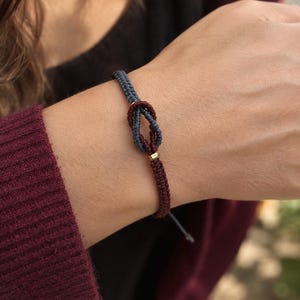 May include: A cord bracelet featuring a two-tone design with a slate blue section and a deep burgundy section, joined by a central knot. The bracelet includes a small, polished gold-toned metal bead accent positioned near the knot.