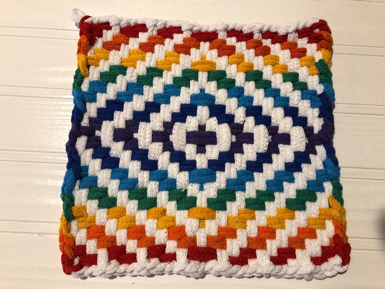 May include: A rainbow-colored woven pot holder with a white background. The pot holder is made of thick cotton yarn and has a checkered pattern.