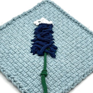 May include: A square, light blue woven potholder with a tree design. The tree is made of dark blue, green, and white knotted fabric. The potholder has a woven border.