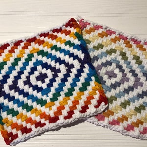 May include: Two colorful woven pot holders with a rainbow pattern. The pot holders are made of thick yarn and have a white background with stripes of red, orange, yellow, green, blue, indigo, and violet. The pot holders are square shaped and have a fringe around the edges.