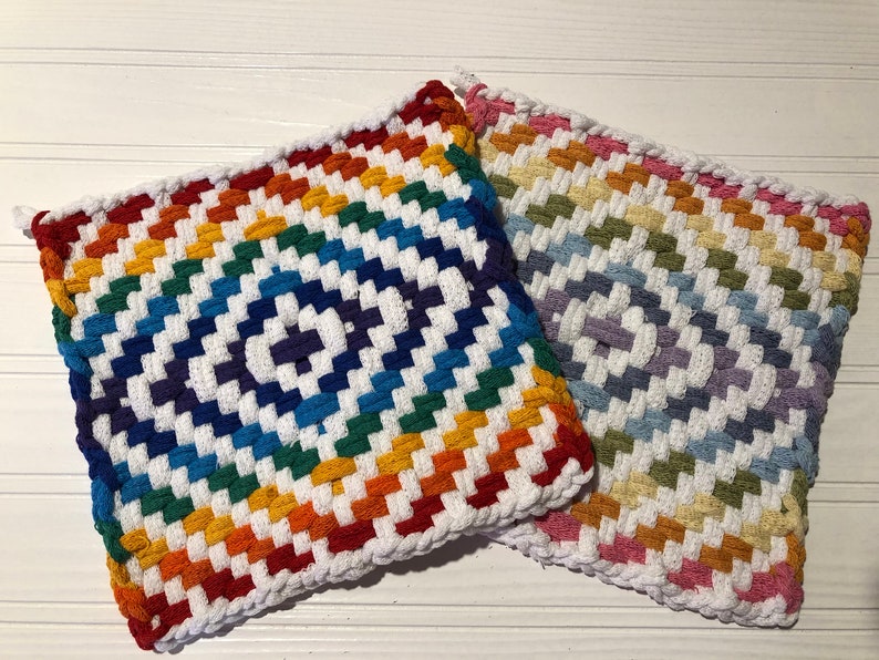 May include: Two colorful woven pot holders with a rainbow pattern. The pot holders are made of thick yarn and have a white background with stripes of red, orange, yellow, green, blue, indigo, and violet. The pot holders are square shaped and have a fringe around the edges.