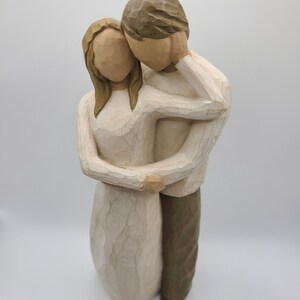 May include: A carved wooden figurine of a couple embracing. The woman wears a cream-colored dress, and the man wears a cream-colored shirt and brown pants. The sculpture has a textured, hand-carved appearance, with a neutral color palette.