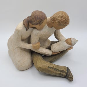 May include: A carved figurine depicting a couple gazing at a newborn baby. The figures are in neutral tones of beige, brown, and tan. The sculpture captures a moment of tenderness and family connection.