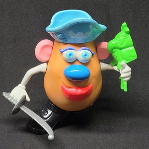 May include: A plastic toy figure of a potato with a blue hat, pink ears, blue eyes, a blue nose, red lips, and a gray sword. The potato is holding a green plastic toy parrot.