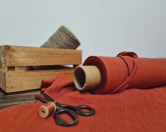 May include: A roll of rust-colored fabric with a cardboard core, partially unrolled on a wooden surface. A wooden crate holds a spool of gray thread. A pair of scissors and a spool of brown thread are also present, suggesting a sewing or crafting theme.