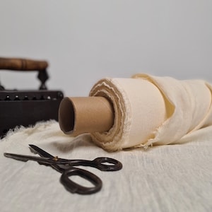 Op de afbeelding: Een rol crèmekleurige stof met een kartonnen kern ligt naast een vintage strijkijzer en een schaar. De stof is uitgerold en toont zijn textuur. Het strijkijzer is zwart met een houten handvat.