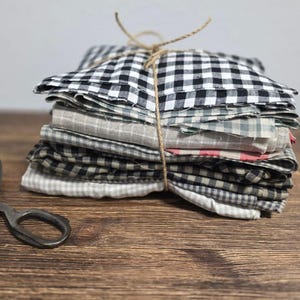 Può includere: Una pila di quadrati di tessuto assortiti, legati con spago. I tessuti presentano vari motivi, tra cui quadretti bianchi e neri, righe e colori uniti. Forbici vintage sono appoggiate su una superficie di legno.