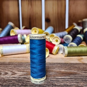 Könnte beinhalten: Eine Spule türkisfarbenes Garn der Marke Gutermann steht vor einer Holzkiste voller verschiedener Garnspulen. Die türkisfarbene Spule ist mit dem Markennamen und 100.6 Metern beschriftet. Weitere Garnfarben sind rot, lila und gelb.