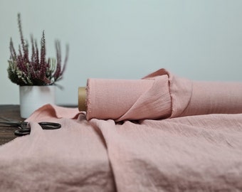 May include: A roll of dusty pink fabric with a visible textured weave, partially unrolled across a surface. A pair of dark metal scissors rests on the fabric near a white pot containing purple and white heather flowers.