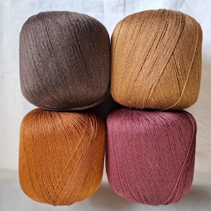 May include: Four balls of yarn arranged in a square formation. The top left ball is dark brown, the top right is golden yellow, the bottom left is burnt orange, and the bottom right is a muted mauve pink. Each ball features a smooth, tightly wound texture.