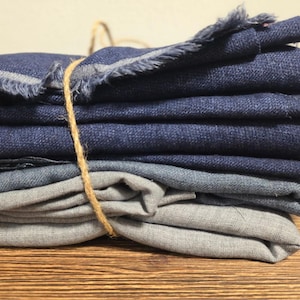 May include: A stack of folded linen napkins in shades of blue and grey, tied with twine. The top napkin has a frayed edge. The napkins are arranged on a wooden surface, creating a rustic aesthetic.
