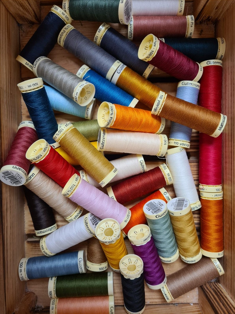 Puede incluir: Una colecci&oacute;n de carretes de hilo de colores, cada uno con una etiqueta que dice "G&uuml;termann" e incluye el texto "100 m" o "110 yds/vgs".