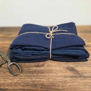 Può includere: Un mucchio di tessuto blu indaco è legato insieme con uno spago. Il tessuto è piegato ordinatamente e si trova su una superficie di legno. Un paio di forbici è posto accanto al tessuto.