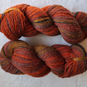 May include: Two skeins of hand-dyed yarn in shades of orange, red, brown, and black. The yarn is tightly wound, showing the texture and colour variations. Suitable for knitting or crochet projects.