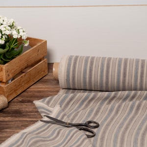May include: A roll of striped fabric in neutral tones of beige, grey, and blue, with a pair of scissors and a length of the fabric laid out. A wooden crate holds a potted plant with white flowers. Twine spools are also present.