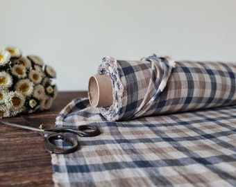 May include: A roll of fabric featuring a blue, beige, and white checkered pattern. The fabric is wrapped around a cardboard tube with frayed edges visible at the end. A pair of metal scissors lies on the fabric surface next to the roll.