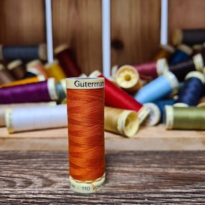 Op de afbeelding: Een spoel oranje garen met de merknaam "Gütermann" gedrukt op het etiket. De spoel is omringd door andere spoelen garen in verschillende kleuren.