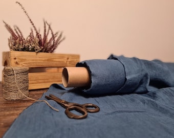 May include: A roll of blue fabric with a cardboard core, scissors, and twine on a wooden surface. A small wooden crate holds dried purple flowers. The scene suggests a craft or sewing project.