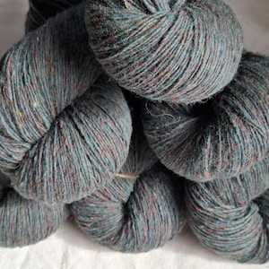 Op de afbeelding: Close-up van verschillende strengen garen in een blauwgroene kleur met spikkels van rood, geel en blauw. Het garen is strak opgerold, wat de textuur van de vezels laat zien. Het garen is geschikt voor brei- of haakprojecten.