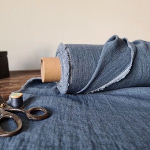 Puede incluir: Un rollo de tela azul con un borde sin rematar, exhibido con herramientas de costura vintage. La tela es de color azul medio y parece ser lino. Tambi&eacute;n se ven unas tijeras, una bobina de hilo y una plancha.
