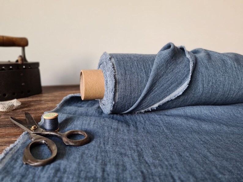 Puede incluir: Un rollo de tela azul con un borde sin rematar, exhibido con herramientas de costura vintage. La tela es de color azul medio y parece ser lino. Tambi&eacute;n se ven unas tijeras, una bobina de hilo y una plancha.